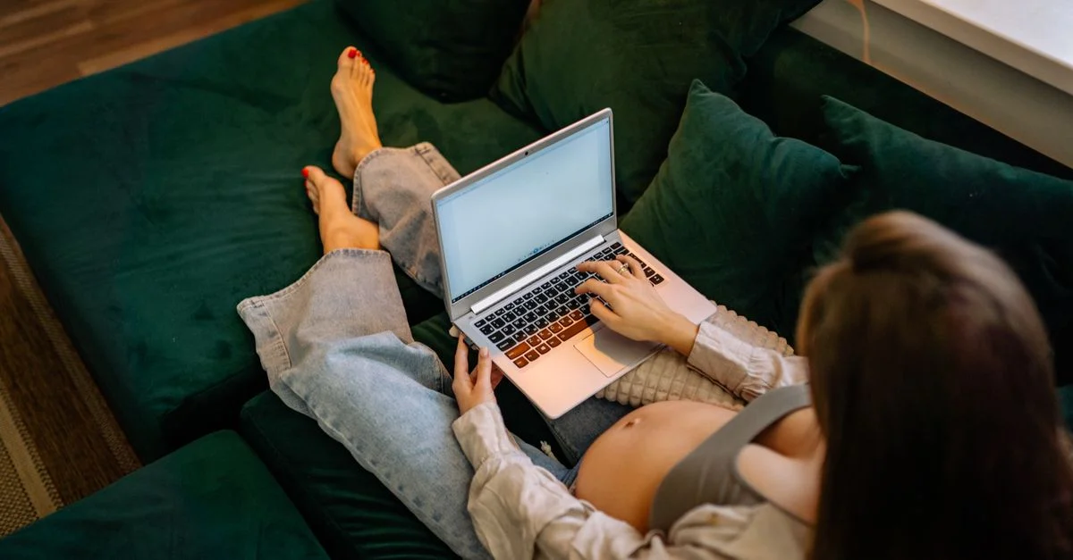 Femme enceinte assise confortablement sur un canapé utilisant un ordinateur portable, travaillant à distance.