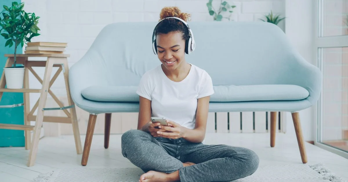 Femme souriante utilisant un smartphone avec des écouteurs dans un salon confortable.