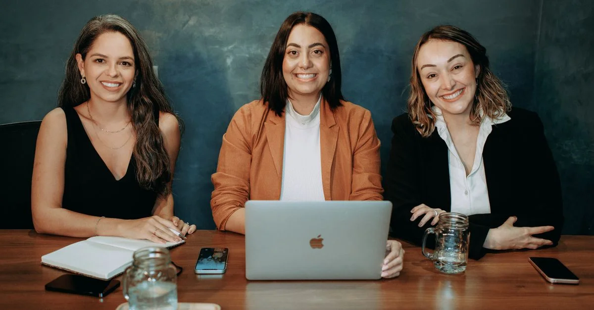 Trois femmes professionnelles assises à un bureau en bois dans un bureau avec des ordinateurs portables et des blocs-notes.